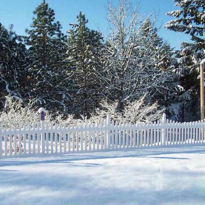 Ellington white vinyl picket fence in snow with trees behind